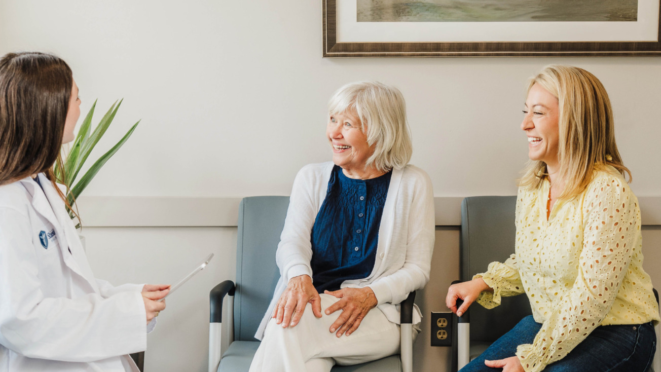 Doctor discussing with two women sitting in chairs, smiling and engaged in conversation in a medical office setting.