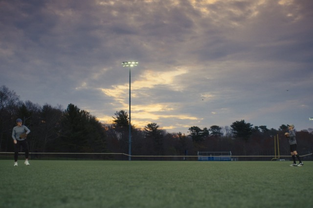 Two people playing catch on a grassy field at dusk with illuminated field lights and a cloudy sky in the background.