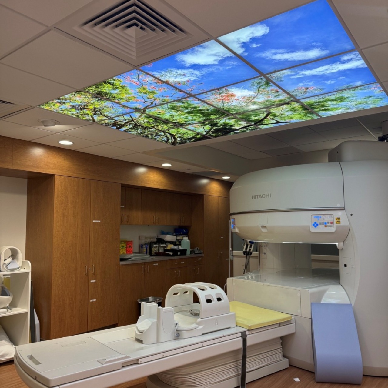 Medical imaging room with MRI machine, ceiling with sky and trees design, and wooden cabinets.