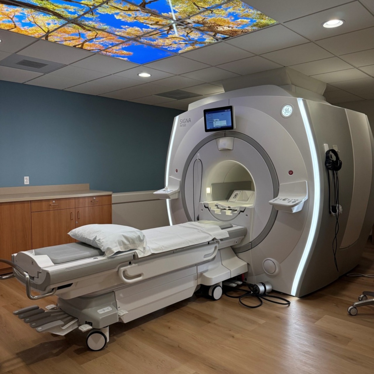MRI machine in a modern medical imaging room with a decorative ceiling panel featuring a tree and blue sky design.