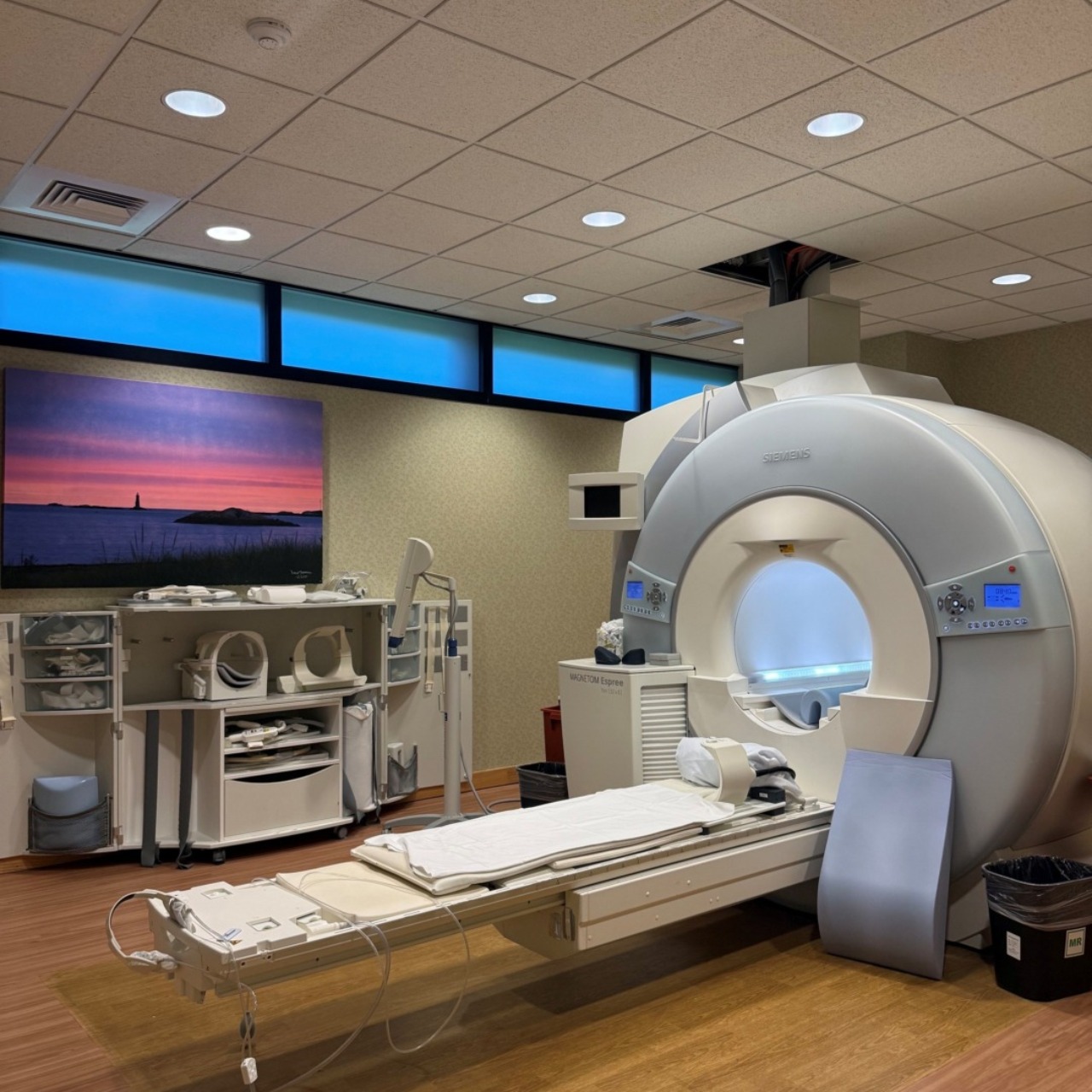 MRI machine in a medical examination room with wooden flooring and a scenic landscape painting on the wall.