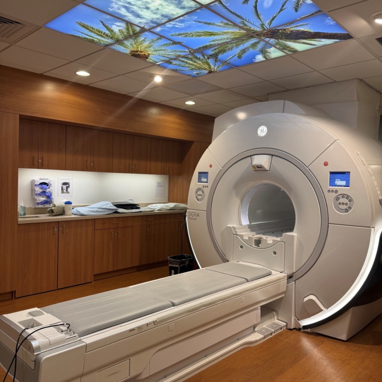 MRI machine in a modern medical room with wooden cabinets and a ceiling light with a tropical sky design.