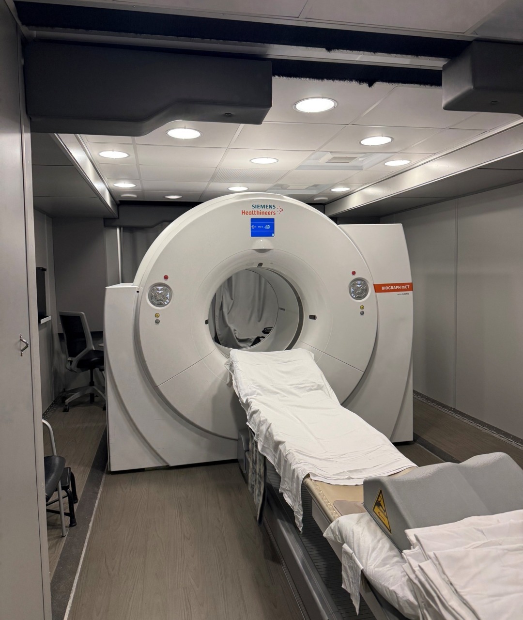 CT scanner in a medical room with a bed positioned for patient imaging, surrounded by modern equipment and lighting.