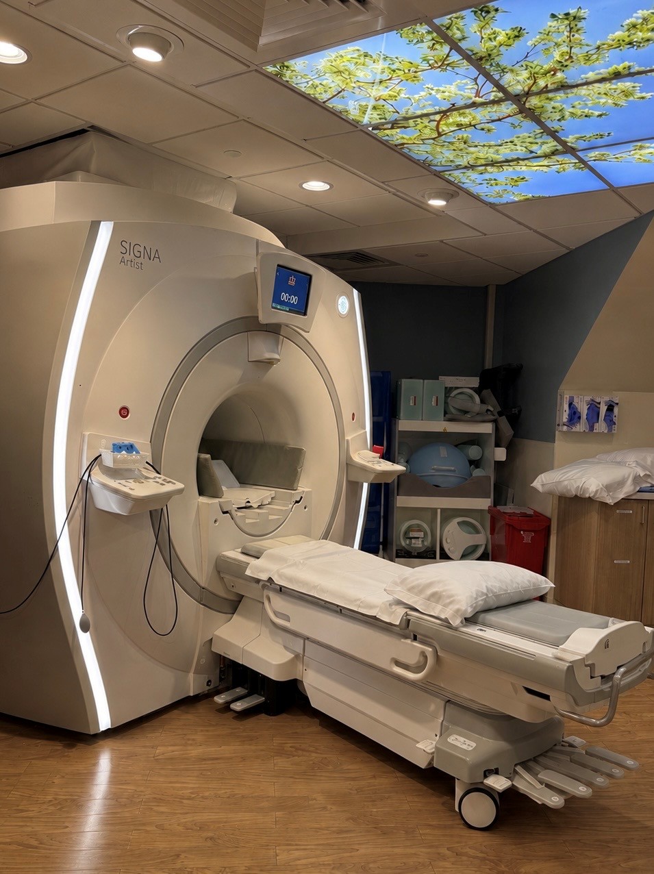 MRI machine in a medical room with a nature-themed ceiling panel and a wooden floor.