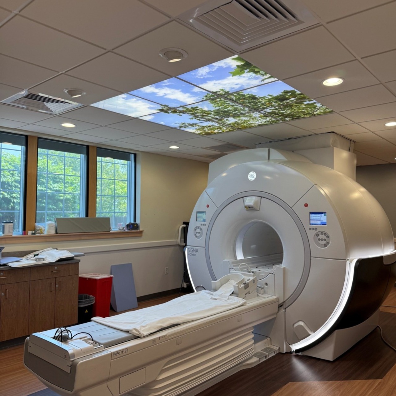 MRI machine in a hospital room with a calming nature-themed ceiling display and large windows.