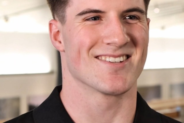Smiling person wearing a black shirt with a beverage brand logo, standing indoors with blurry background.