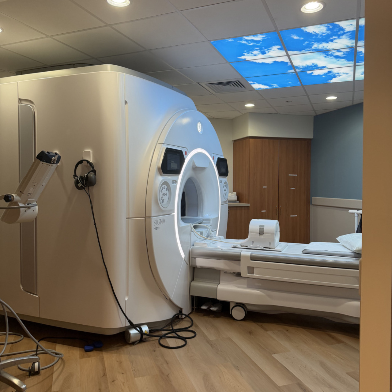 MRI machine in a hospital room with a ceiling light panel displaying blue sky and clouds, wooden accents, and modern design.