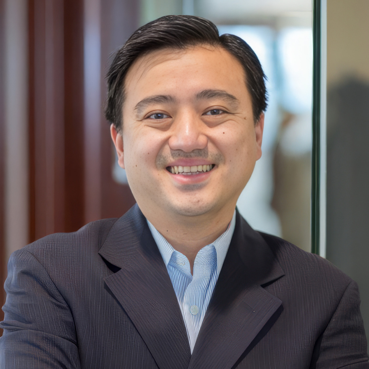 Smiling person in a suit standing indoors with arms crossed, blurred background.