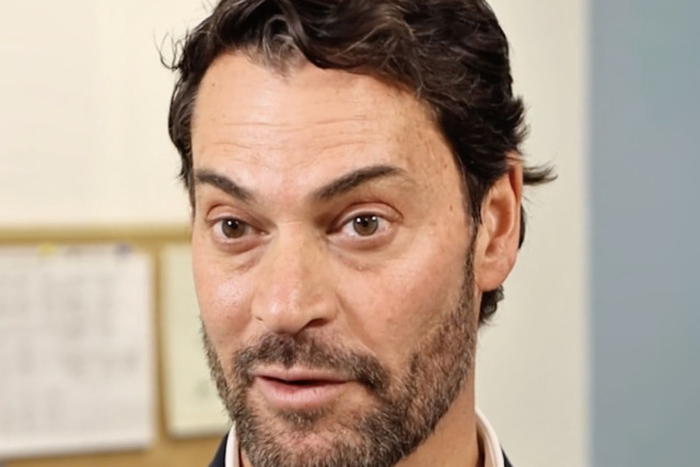 Man with short dark hair and beard wearing a suit, speaking indoors with a blurred bulletin board in the background.